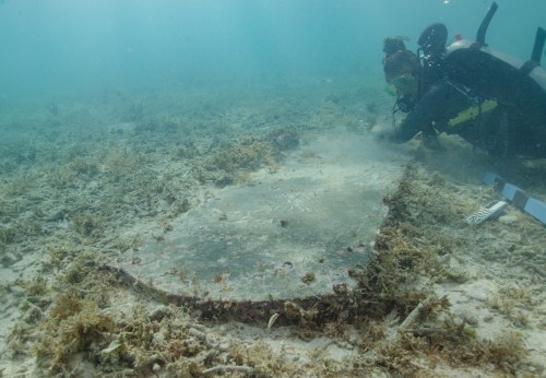 A diver examines a large, submerged object on the ocean floor, surrounded by seaweed and marine life.
