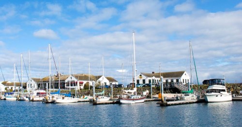 A scenic harbor with various sailboats and yachts docked alongside a charming waterfront building under a blue sky.