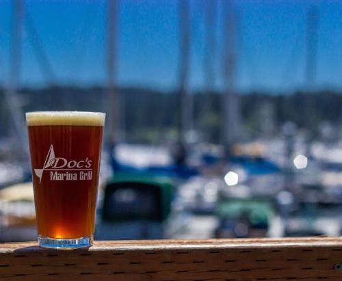 A pint of beer sits on a wooden railing with a blurred marina in the background.