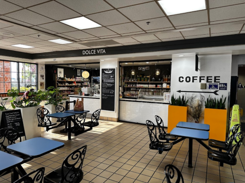 A cozy café interior with blue tables, plants, and a counter displaying coffee and pastries. Sign reads "Dolce Vita."