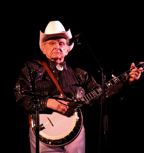 An elderly musician in a cowboy hat plays the banjo on stage, wearing a sparkly black shirt.