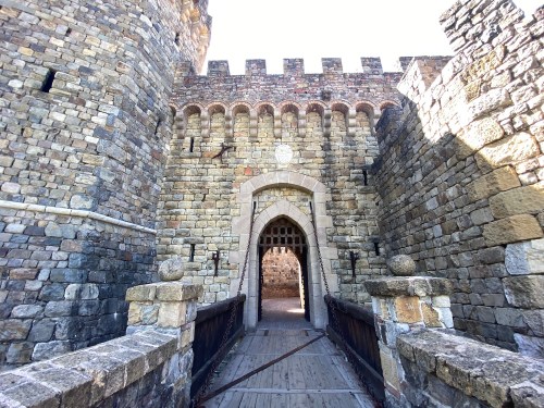 Stone castle entrance with a wooden drawbridge and tall, fortified walls under a clear sky.
