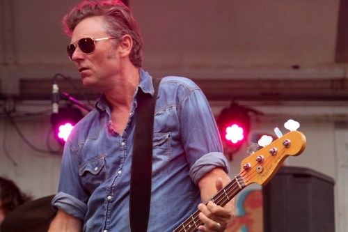 A musician in sunglasses plays bass on stage, wearing a denim shirt, with colorful lights in the background.