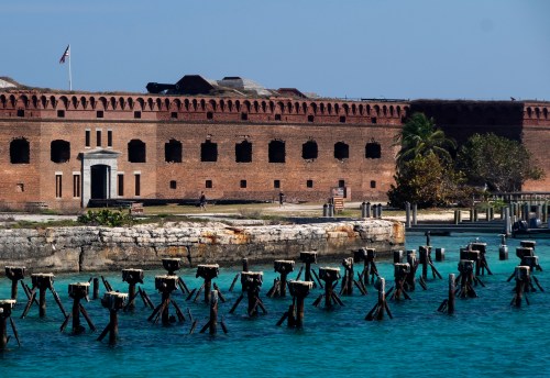 Historic brick fort with arched windows beside turquoise water and wooden pilings, under a clear blue sky.