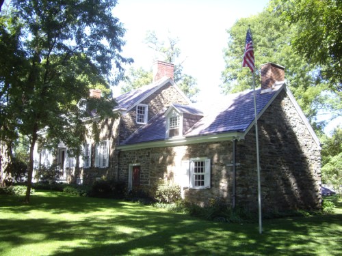 A stone house with a slate roof, surrounded by trees and a well-maintained lawn, featuring an American flag.