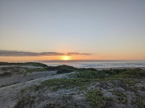 A serene beach scene at sunset, with gentle waves and grassy dunes under a colorful sky.