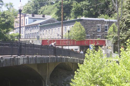 A stone bridge leads to a sign reading "Ellicott City," surrounded by greenery and historic buildings.