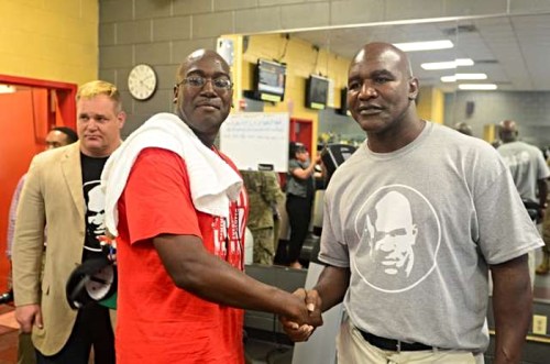 Two men shake hands in a gym, one wearing a red shirt and the other in a gray t-shirt, with a third person in the background.