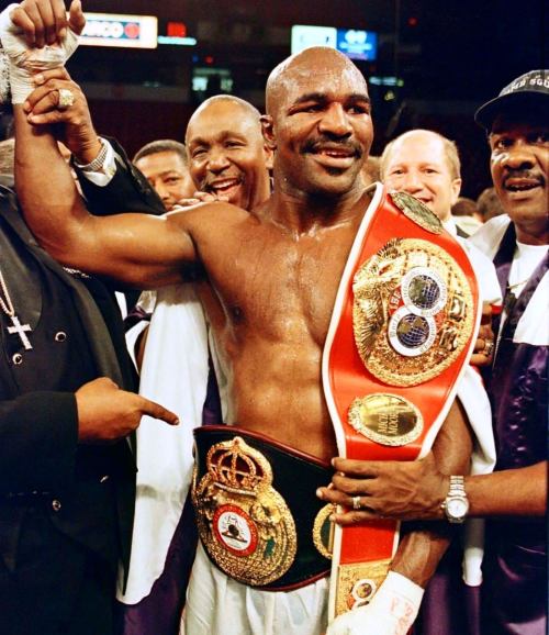 A victorious boxer holds up his championship belts, surrounded by smiling supporters in a celebratory atmosphere.