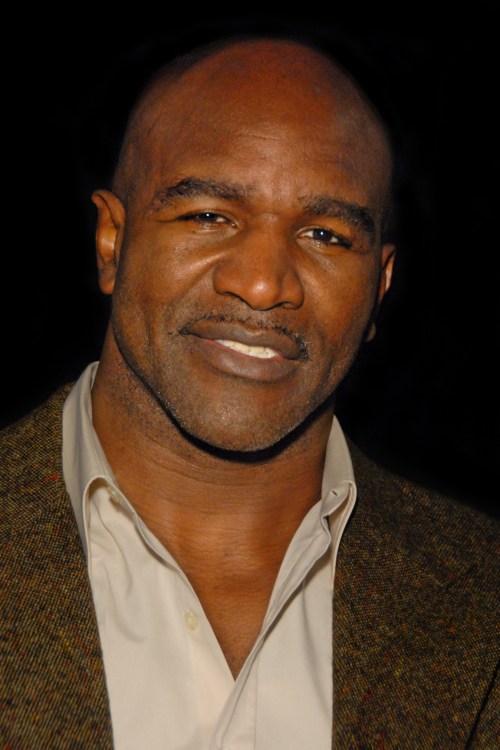 A smiling man with a bald head, wearing a light shirt and a dark blazer, poses for the camera against a dark background.