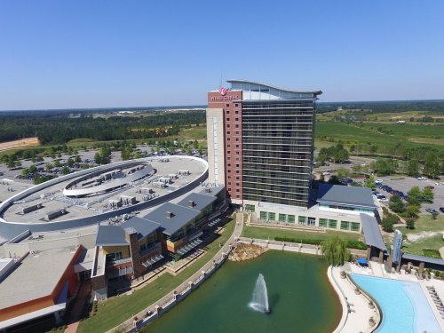 Aerial view of a large hotel with a modern design, surrounded by a pond and green landscape under a clear blue sky.
