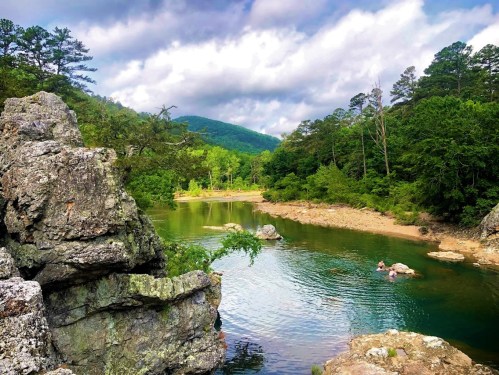 A serene river scene surrounded by lush greenery and rocky formations under a cloudy sky.