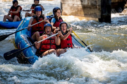 A group of four young women joyfully paddles a blue raft through whitewater rapids, wearing helmets and life jackets.