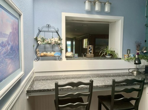 A cozy kitchen nook with a granite countertop, two black chairs, and a decorative shelf with plants and baked goods.