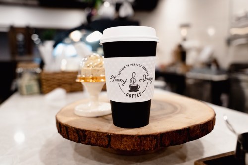 A black coffee cup with a white label reading "Ebony & Ivory Coffee" sits on a wooden surface in a café.