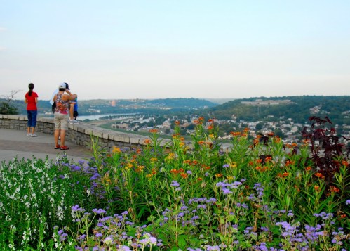 A scenic overlook with colorful flowers in the foreground and people enjoying the view of a river and hills in the background.