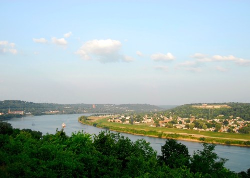 A scenic view of a river winding through green hills and a small town under a clear blue sky with scattered clouds.