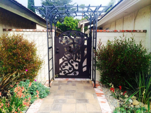 A decorative metal gate with leaf patterns opens to a garden path lined with colorful plants and stones.