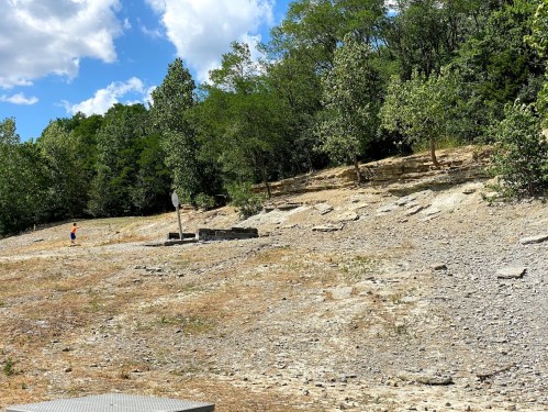 A child walks along a rocky, dry landscape with sparse trees under a partly cloudy sky.