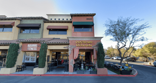 Exterior view of a building featuring a pasta restaurant and a fitness center, with outdoor seating and greenery.