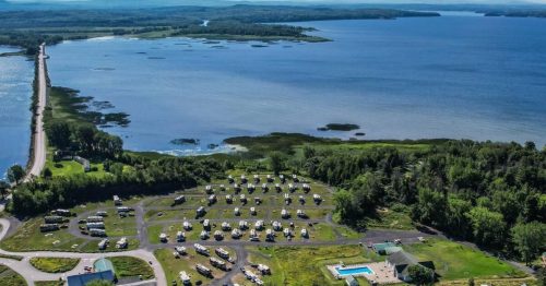 Aerial view of a lakeside RV park with numerous campers, a pool, and a scenic waterway in the background.