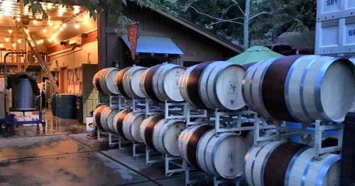 A row of wine barrels outside a rustic winery, with warm lights glowing in the background.