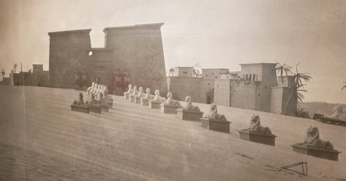Black and white photo of ancient Egyptian architecture with sphinx statues and palm trees in a desert landscape.