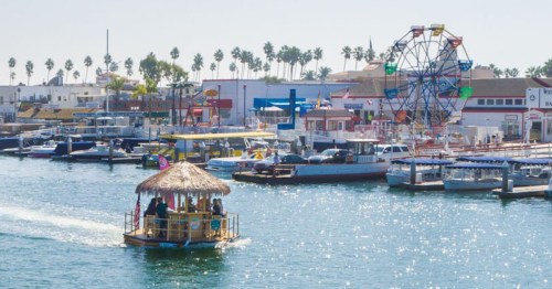 A colorful boat with a thatched roof navigates a sunny harbor filled with boats and a ferris wheel in the background.