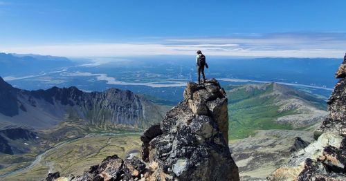 A hiker stands on a rocky peak, overlooking a vast landscape of mountains and a river below under a clear blue sky.