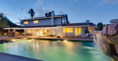 A modern home with a pool and waterfall, surrounded by palm trees, under a twilight sky.