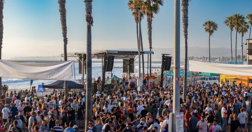 A large crowd gathers at a beach event with palm trees, a stage, and food trucks in the background.