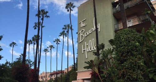 Sign for "The Beverly Hills" surrounded by palm trees and greenery under a blue sky with scattered clouds.