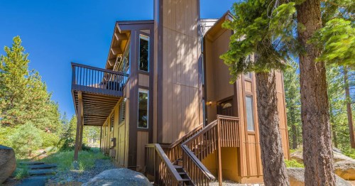 A modern wooden house surrounded by trees, featuring a deck and multiple windows under a clear blue sky.