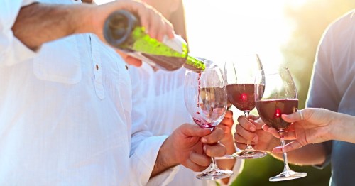 A group of people toasting with wine glasses as one pours red wine into a glass, with sunlight in the background.