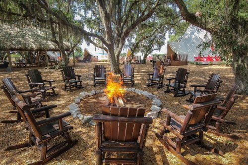 A circle of wooden chairs around a campfire, surrounded by trees and tents in a rustic outdoor setting.