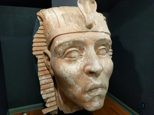A large, ancient stone sculpture of a pharaoh's head, partially damaged, displayed against a dark background.