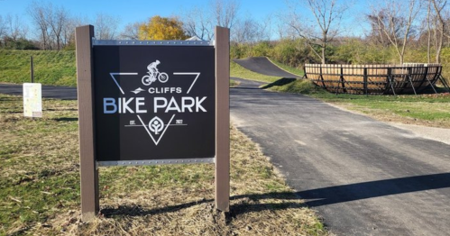 Sign for "Cliffs Bike Park" with a bike graphic, surrounded by grassy areas and a bike ramp in the background.