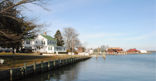 A serene waterfront scene featuring white houses, trees, and a dock along a calm river under a clear blue sky.