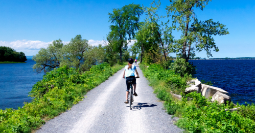 A person rides a bicycle along a scenic path by a river, surrounded by greenery and blue skies.