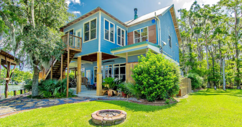 A blue two-story house surrounded by greenery, featuring a fire pit and a staircase leading to a deck.