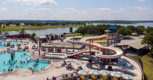 Aerial view of a busy water park with a large slide, swimming pool, and people enjoying the sunny day by the water.