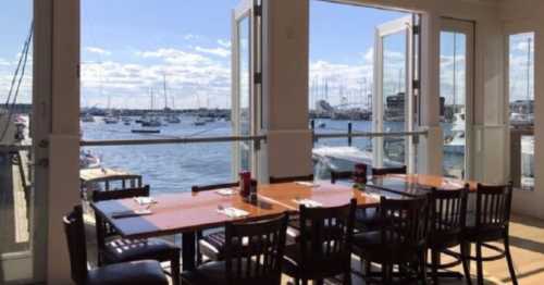 A waterfront restaurant with open doors, featuring a table set for dining and boats in the harbor outside.