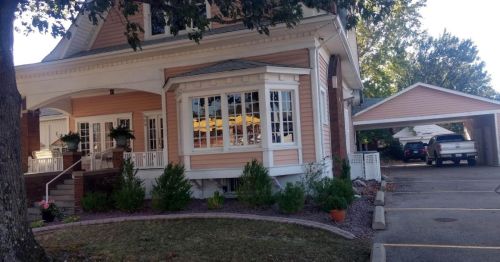 A charming pink house with a porch, surrounded by greenery, and a carport nearby.