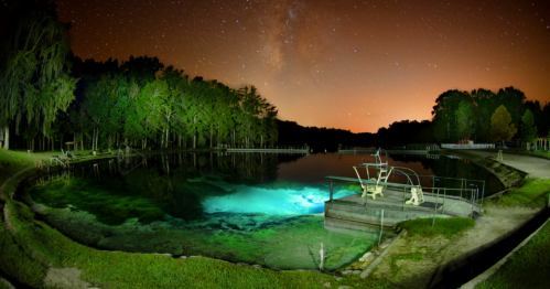 A serene night scene of a lake surrounded by trees, illuminated by stars and a glowing underwater spring.