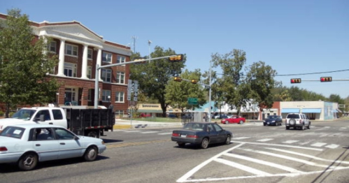 Traffic at an intersection with a historic building, trees, and various vehicles on a sunny day.