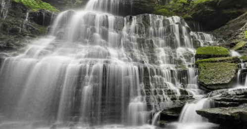 A cascading waterfall flows over rocky cliffs, surrounded by lush greenery and moss.