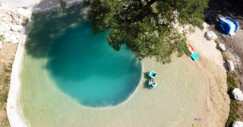 Aerial view of a circular, clear blue swimming area surrounded by sandy beach and greenery. Two people float on rafts.