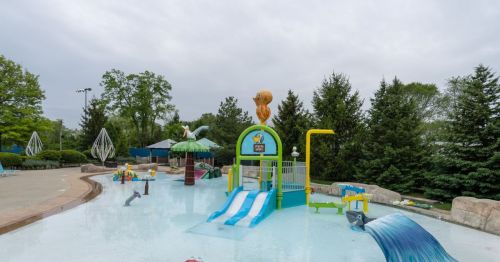 A colorful water play area with slides, fountains, and playful structures surrounded by greenery under a cloudy sky.