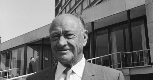 A smiling older man in a suit stands outside a modern building, with another person visible in the background.