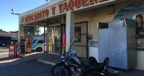 A small market and taqueria with a motorcycle parked outside, featuring a colorful sign and a food cart.